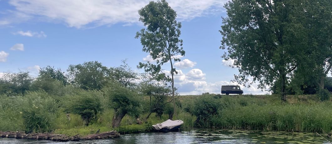 Een auto rijdt op de Lingedijk, terwijl de wind door de bomen waait en de Blokhutboot rustig doorvaart
